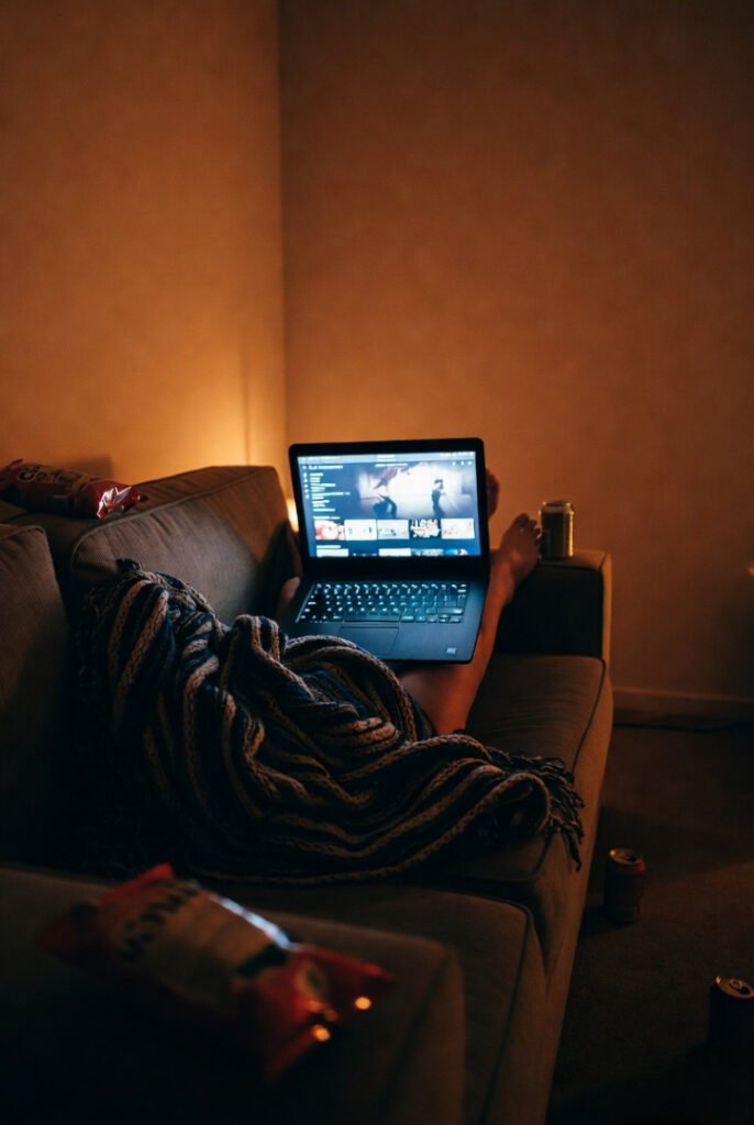 Low-light couch potato life — legs under blanket, streaming on lap, snack wreckage included