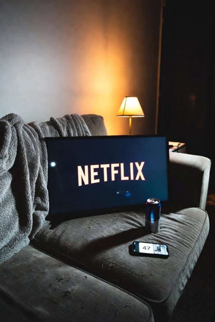 Messy gray couch with fallen fuzzy blanket, paused Netflix screen, empty energy drink can, phone showing 47 notifications, dim sad-cozy lamp glow