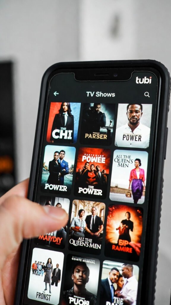 Phone held in hand displaying Tubi’s TV Shows page: grid of show posters, thumb in frame ready to select