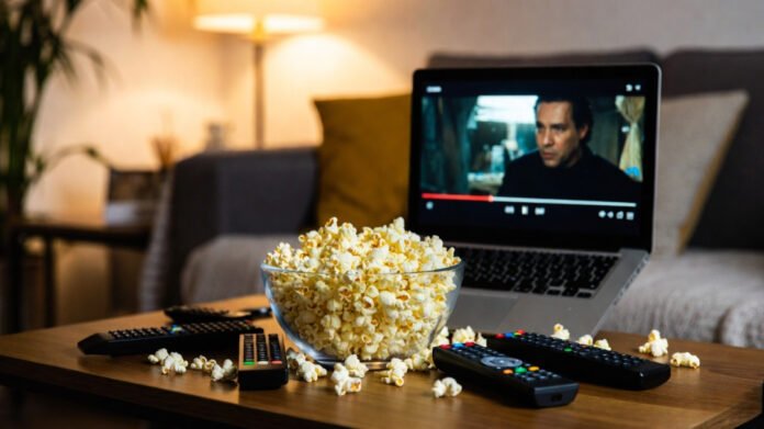 Cozy living room binge setup: popcorn bowl, tangled remotes, laptop on a pillow showing a frozen movie scene.